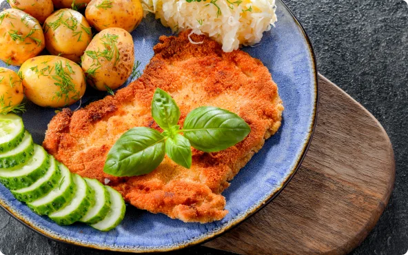 Panierowany kotlet z ziemniakami, kiszoną kapustą i ogórkiem na talerzu.